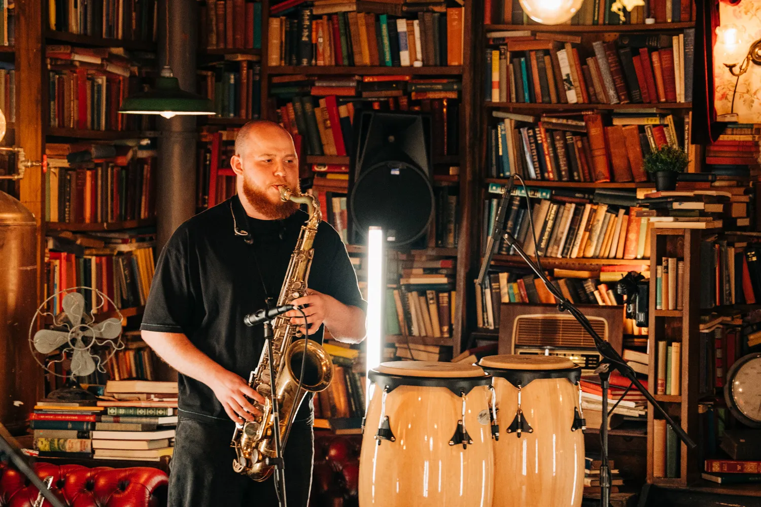 Bearded man playing saxophone in a cozy room filled with books and musical instruments including conga drums.