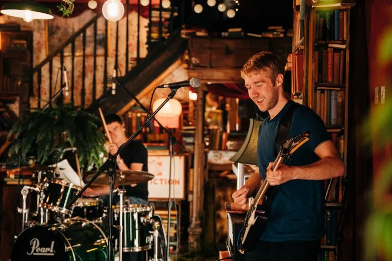 Two musicians playing in a cozy room; one is playing electric guitar and smiling, the other is playing drums in the background.