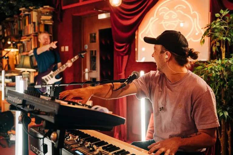 Musician playing keyboard and singing into a microphone with bassist pointing in the background in a cozy, warmly lit room.