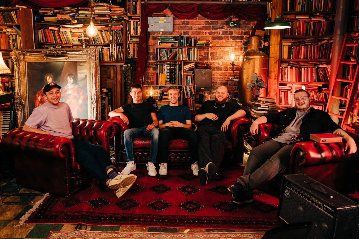 Five men sitting on red leather sofas in a cozy, vintage library filled with books and warm lighting.